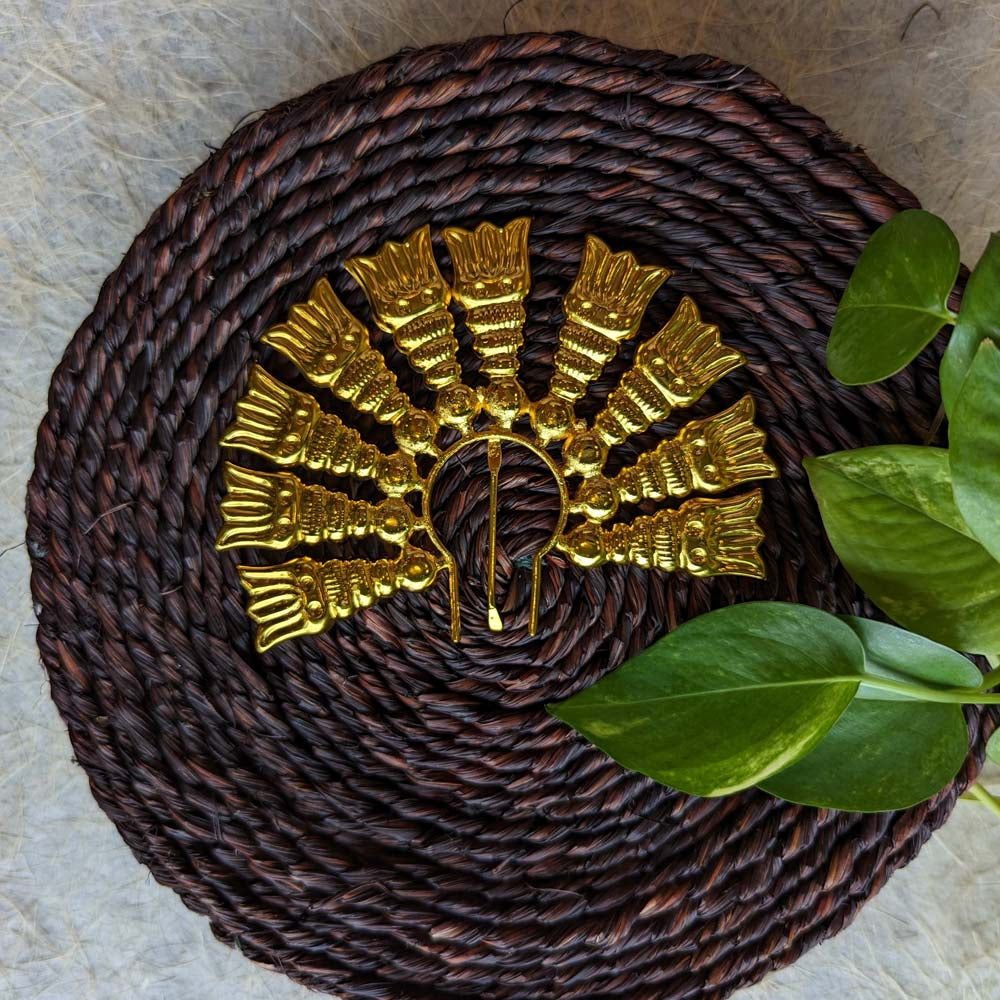 Gold-toned Jada Billai with intricate temple-inspired design, displayed on a woven brown mat with green leaves.