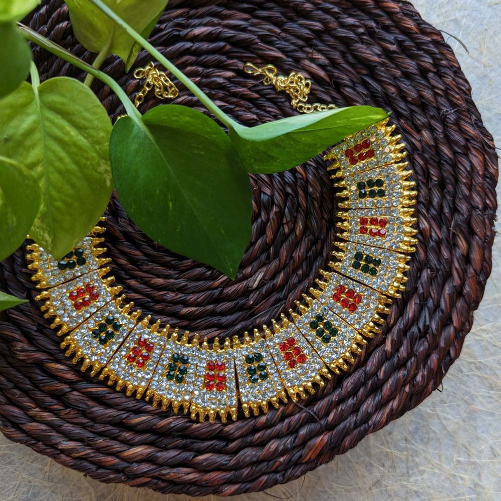 Gold-toned short necklace with ruby and emerald accents and sparkling crystal stones, displayed on a woven brown mat with green leaves.