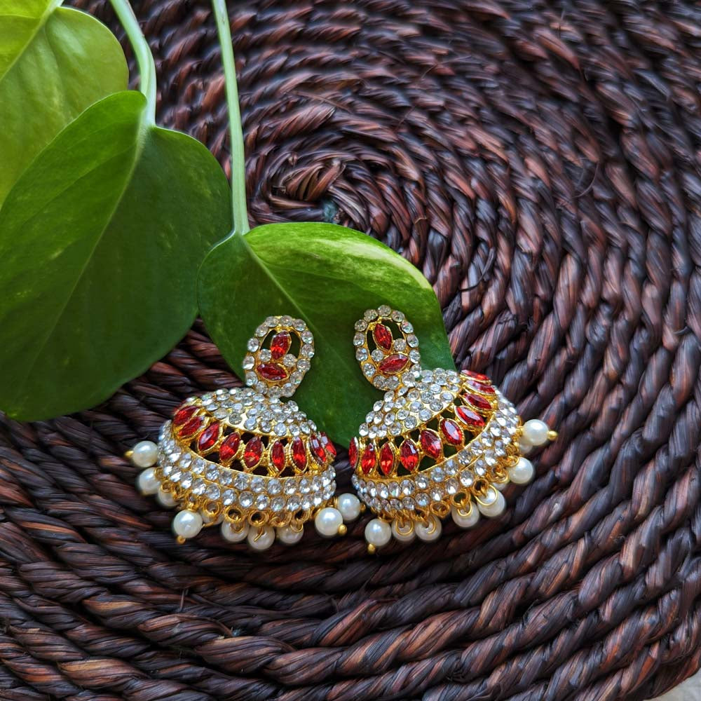 Gold-plated Jhumki earrings with ruby stones, sparkling crystals, and pearl drops, displayed on a woven brown mat with green leaves.