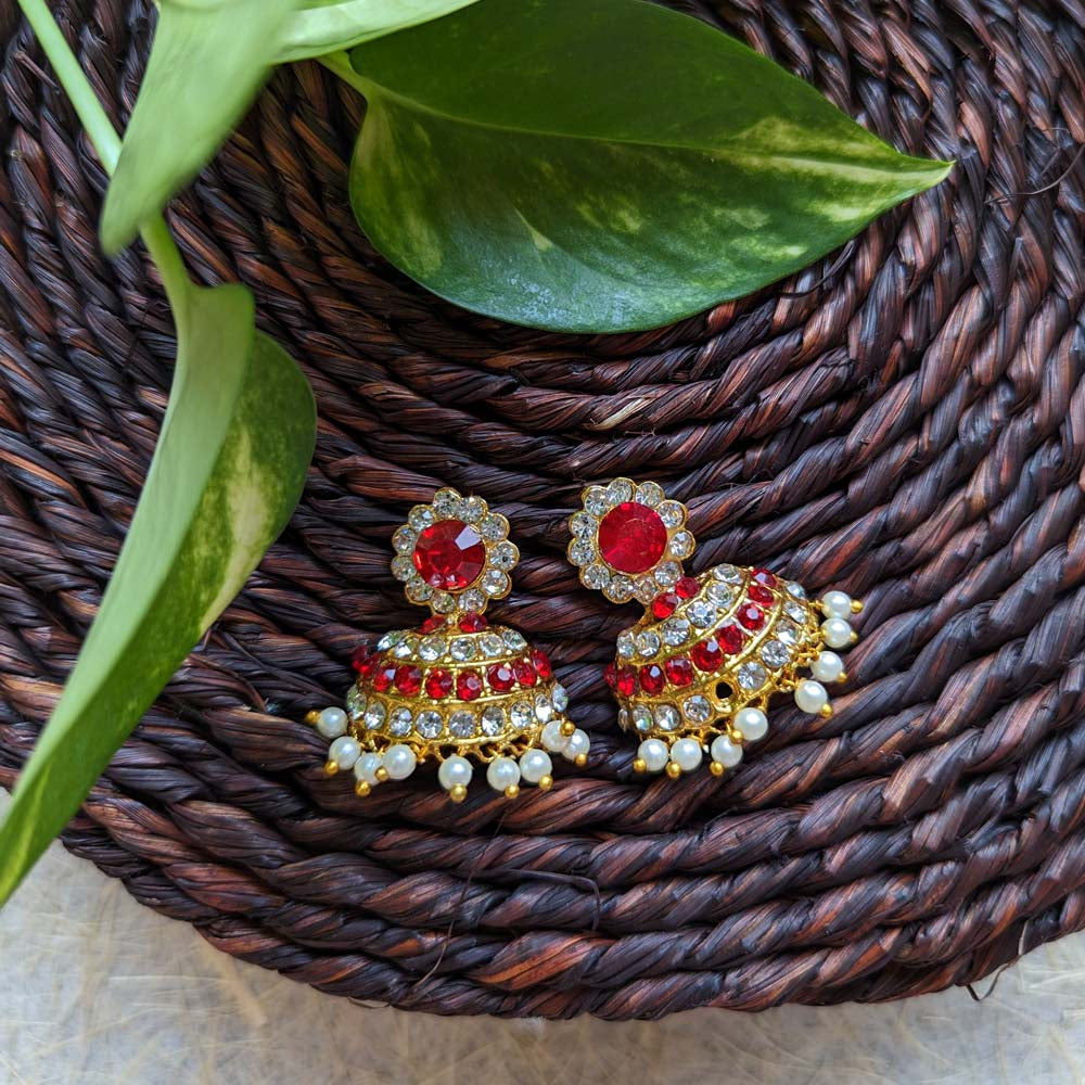 Gold-toned Jhumki earrings adorned with ruby stones, crystals, and pearl drops, displayed on a woven brown mat with green leaves.