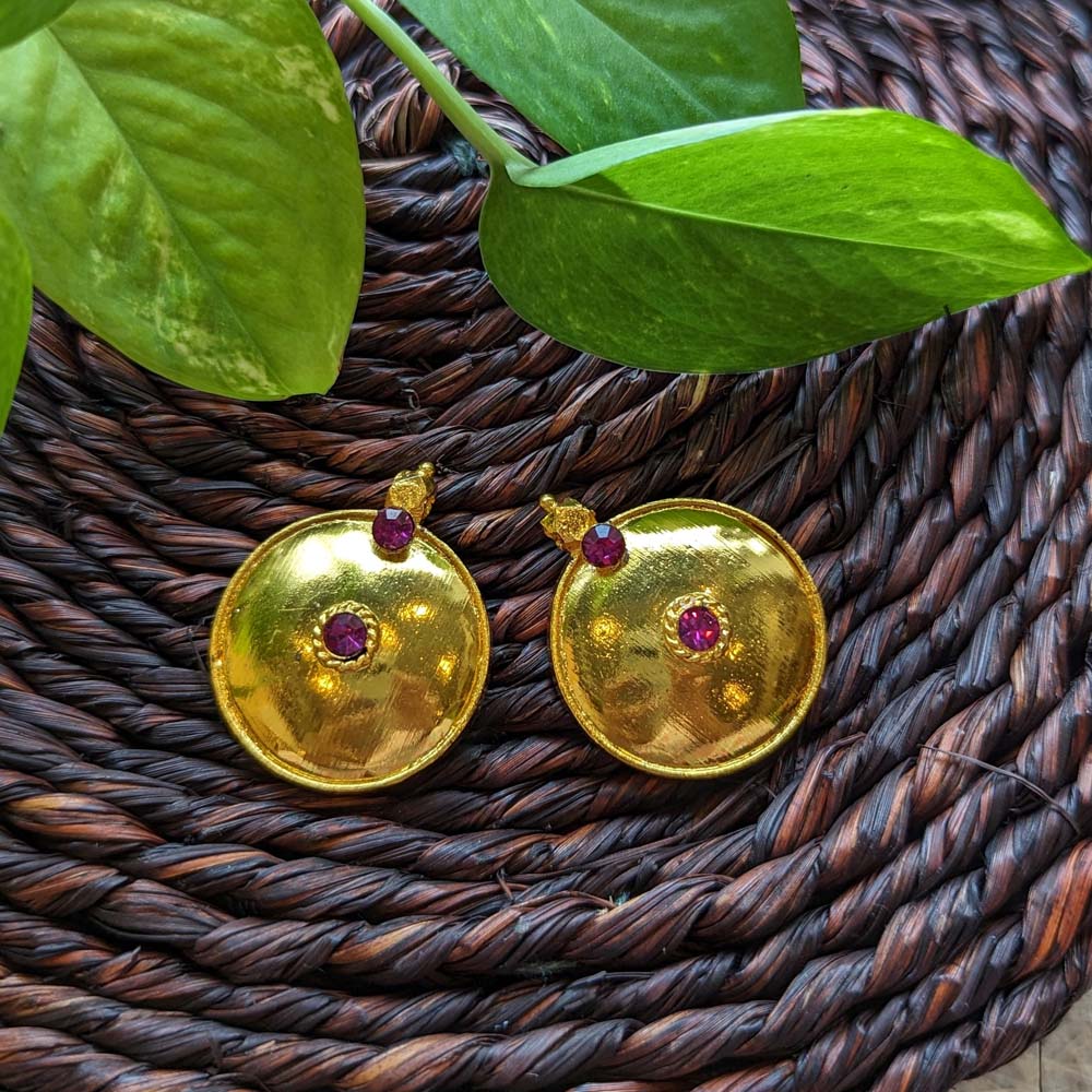 Small traditional gold-plated thali earrings with ruby stone accents, displayed on a woven brown background with green leaves.