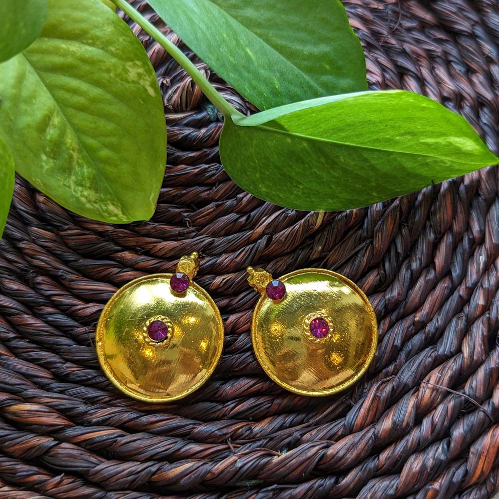 Large traditional gold-plated thali earrings with ruby stone accents, displayed on a woven brown background with green leaves.