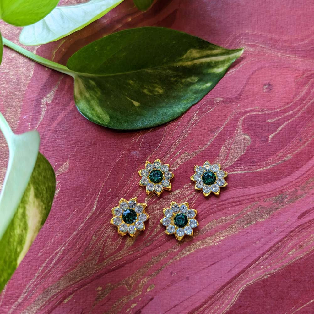Floral decorative stone jewels with gold-toned base, crystal accents, and green centerpieces, displayed on a maroon background with green leaves.