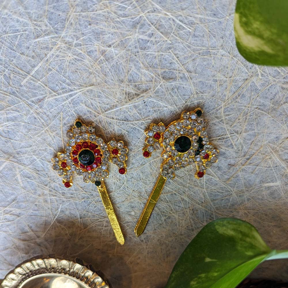 Gold-toned Sankha Chakra ornaments adorned with ruby and emerald stones and sparkling crystals, displayed on a textured background with green leaves.