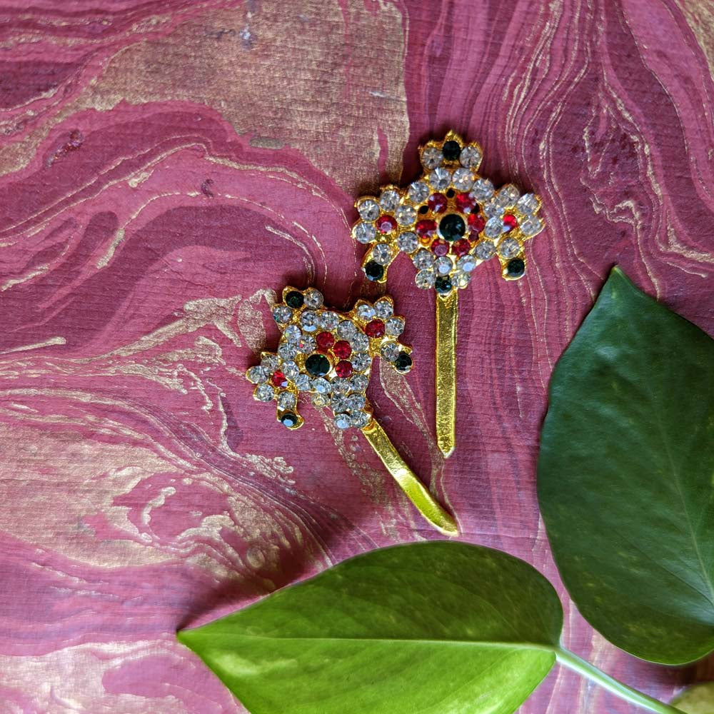 Traditional Sankha Chakra decorative pieces with crystal rhinestones, ruby-red, and emerald-green accents on a gold-toned base, displayed on a maroon background with green leaves.