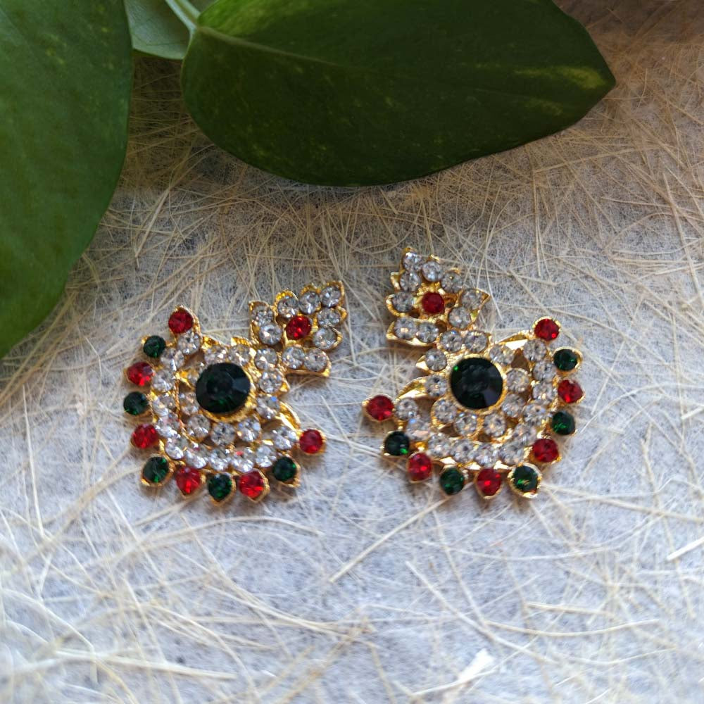 Gold-toned stud earrings with crystal, ruby, and emerald embellishments, displayed on a textured background with green leaves.