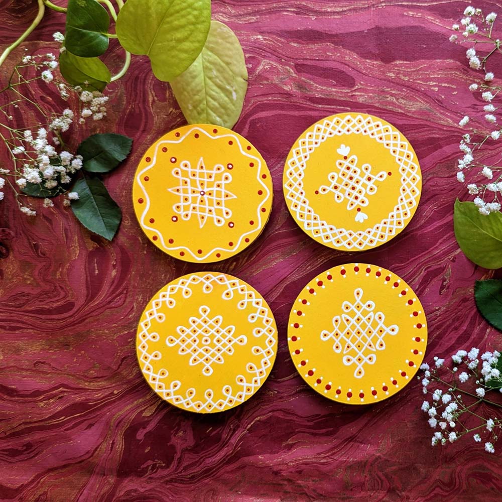 Handcrafted yellow round wooden coasters featuring central kolam designs with intricate white and red patterns, placed on a vibrant decorative background with floral accents.