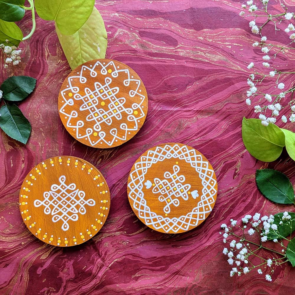 Handcrafted natural brown round wooden coasters with white kolam designs and yellow accents, displayed on a vibrant decorative background with floral embellishments.