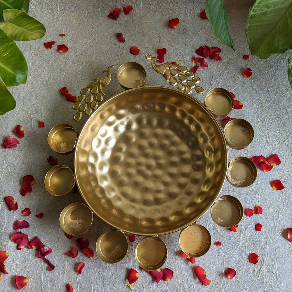 Handcrafted metal urli bowl with hammered golden finish, intricate peacock embellishments, and diya holders.