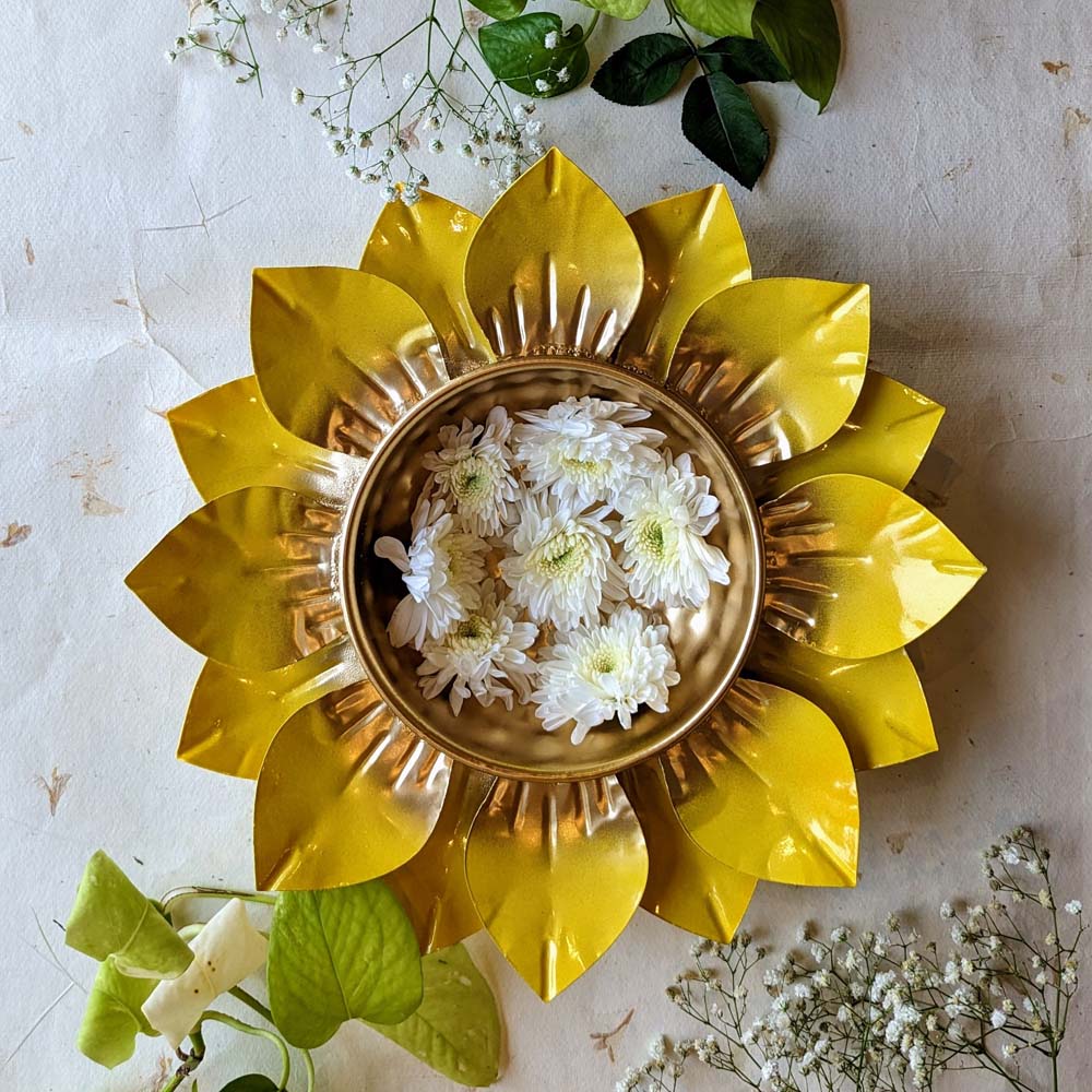 Handcrafted lotus-shaped metal urli bowl with a golden finish and floating white flowers, surrounded by green leaves.