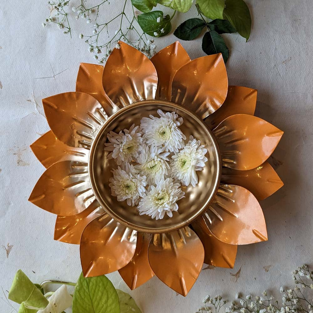 Handcrafted lotus-shaped metal urli bowl with a golden finish and floating white flowers, surrounded by green leaves.