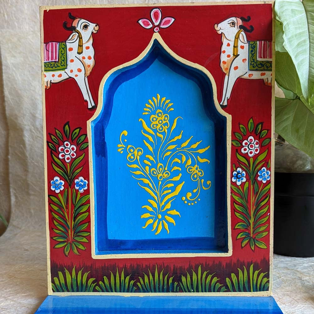 Hand-painted wooden wall shelf in red and blue tones, featuring traditional cow illustrations, floral patterns, and an arched design.