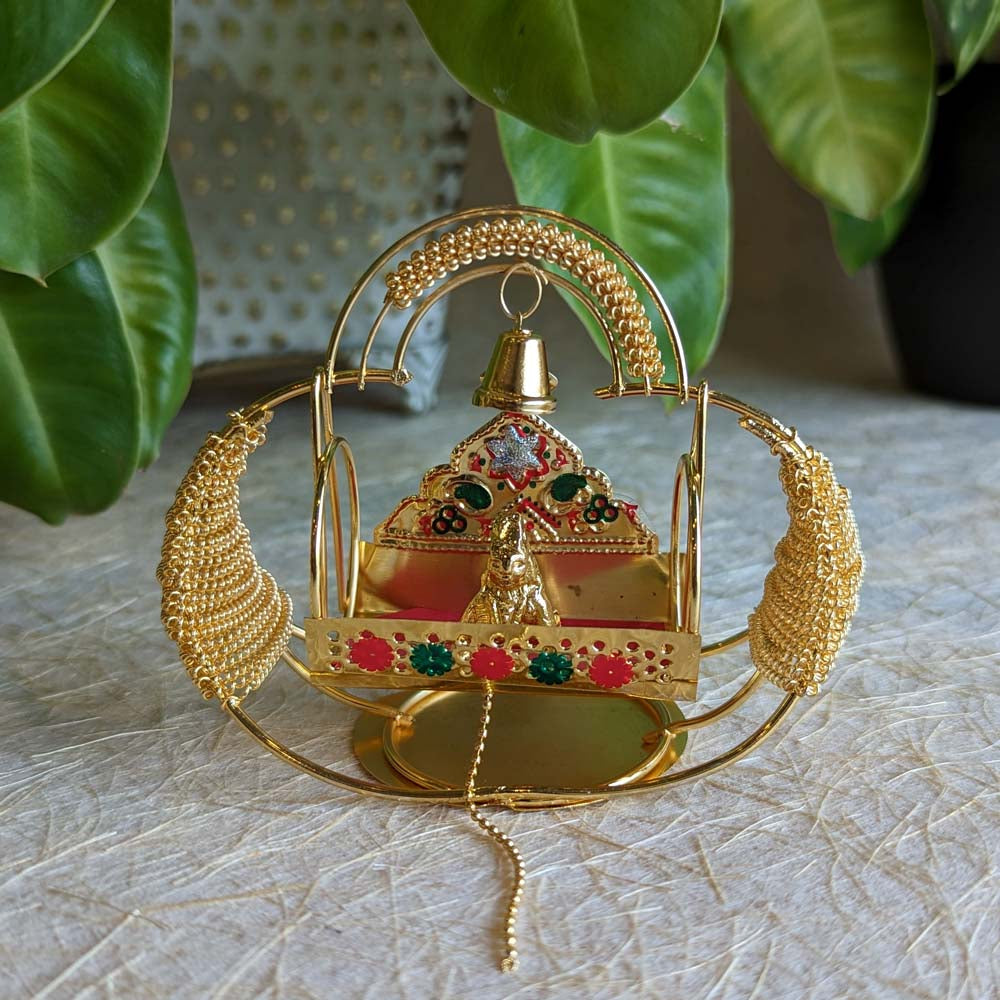 Handcrafted Krishna idol in a golden cradle with colorful accents and a hanging bell, surrounded by decorative elements.