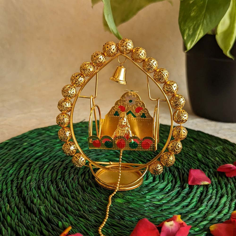 Handcrafted Krishna idol in a golden cradle with colorful accents and a hanging bell, surrounded by decorative elements.
