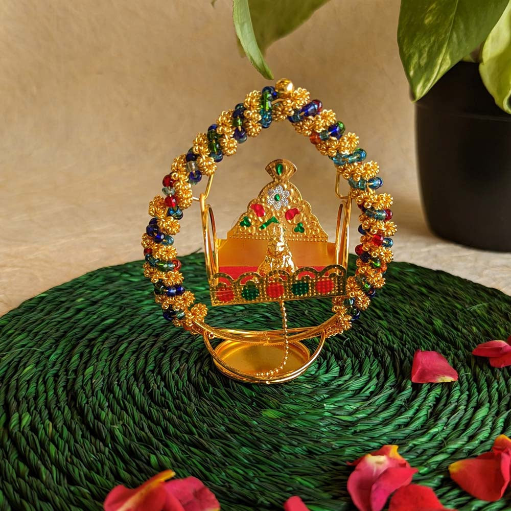 Handcrafted Krishna idol in a golden cradle with colorful accents and a hanging bell, surrounded by decorative elements.