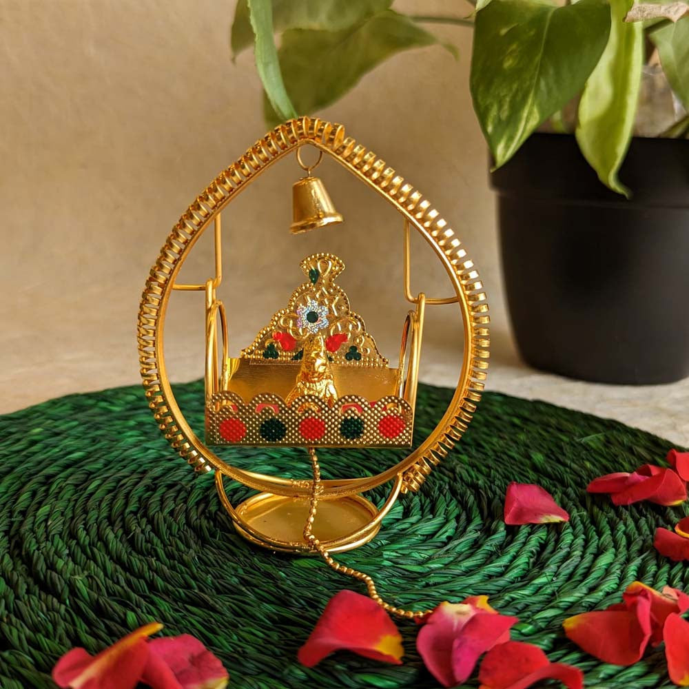 Handcrafted Krishna idol in a golden cradle with colorful accents and a hanging bell, surrounded by decorative elements.