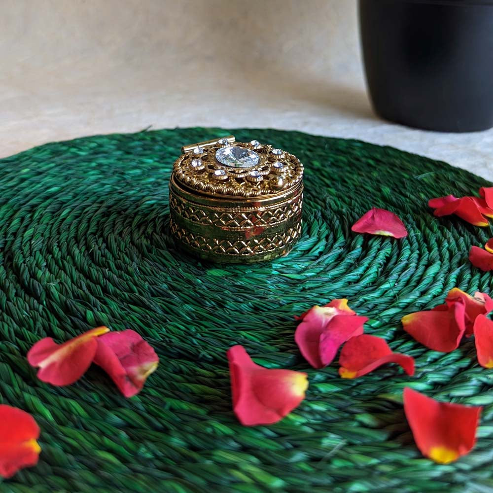 Golden round kumkum box with gemstone embellishments, placed on a green woven mat with scattered rose petals.