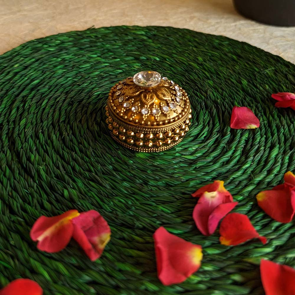 Small round kumkum box in gold with crystal accent, placed on a green woven mat surrounded by rose petals.