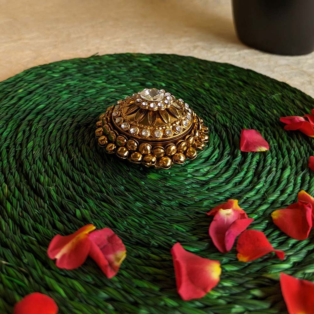 Round kumkum box in gold with ornate design and crystal accents, placed on a green woven mat with scattered rose petals.