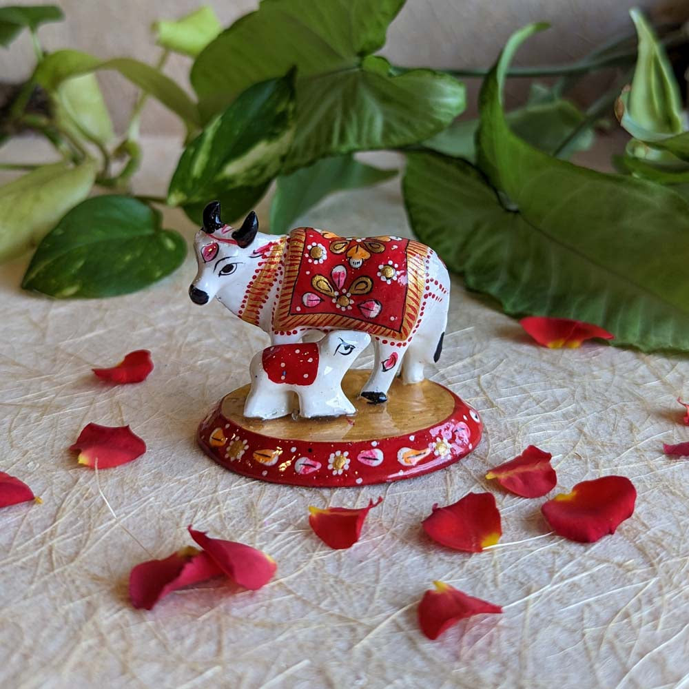 Handcrafted Kamadhenu and calf figurine with vibrant red, white, and gold detailing, surrounded by rose petals and green foliage.