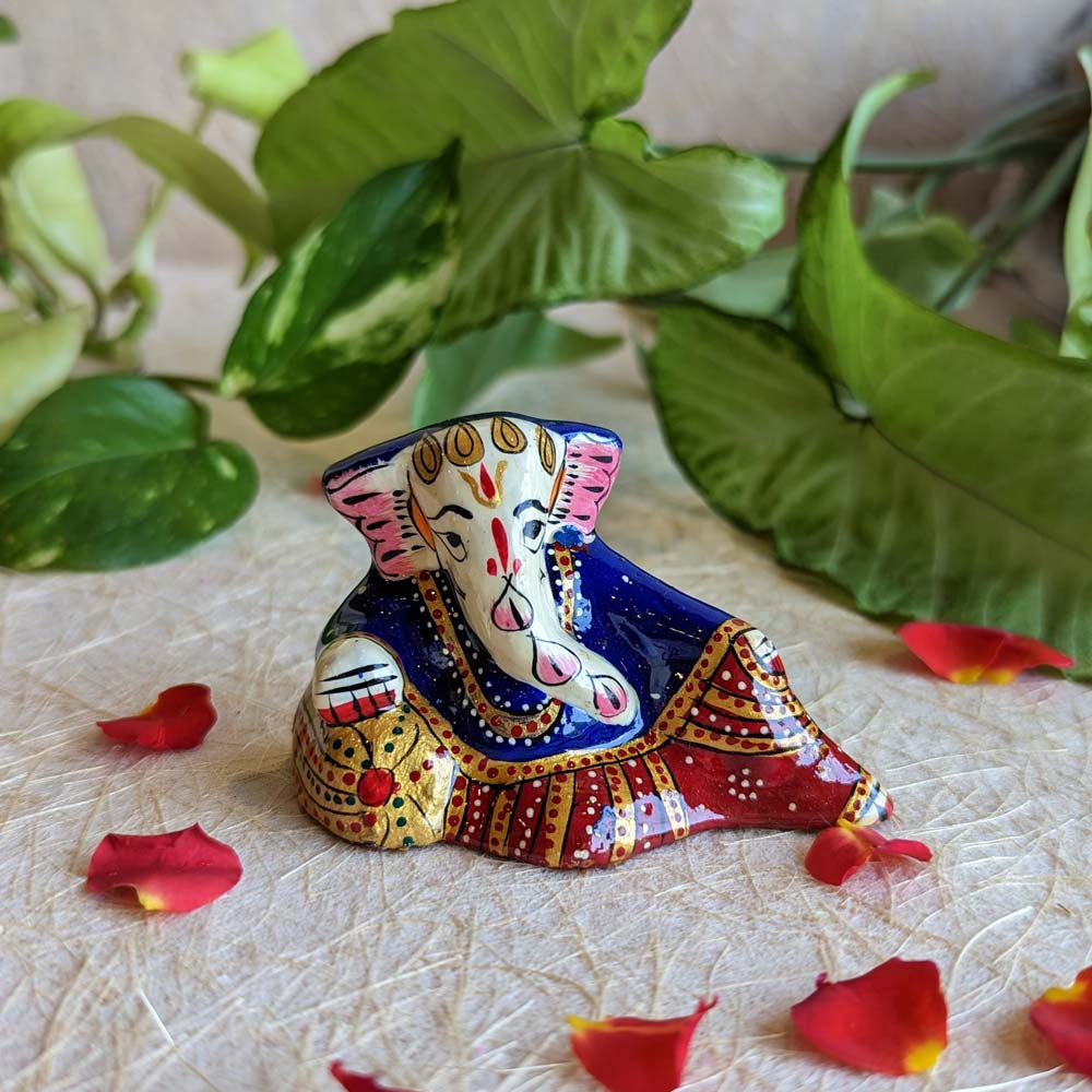 Handcrafted Reclining Ganesha figurine with vibrant blue, red, and gold detailing surrounded by rose petals and green foliage.