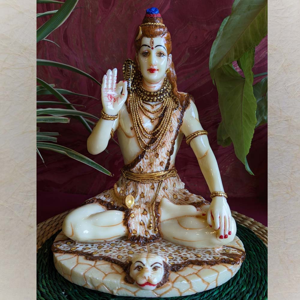 Handcrafted Blessing Shiva idol in ivory and gold finish, seated in a meditative pose on a tiger skin, with one hand raised in blessing, displayed on a green mat with a red backdrop and foliage.