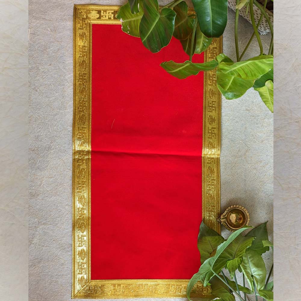 A vibrant red Aasan with an intricate gold border, displayed on a textured surface with green leaves and a brass diya for decor.
