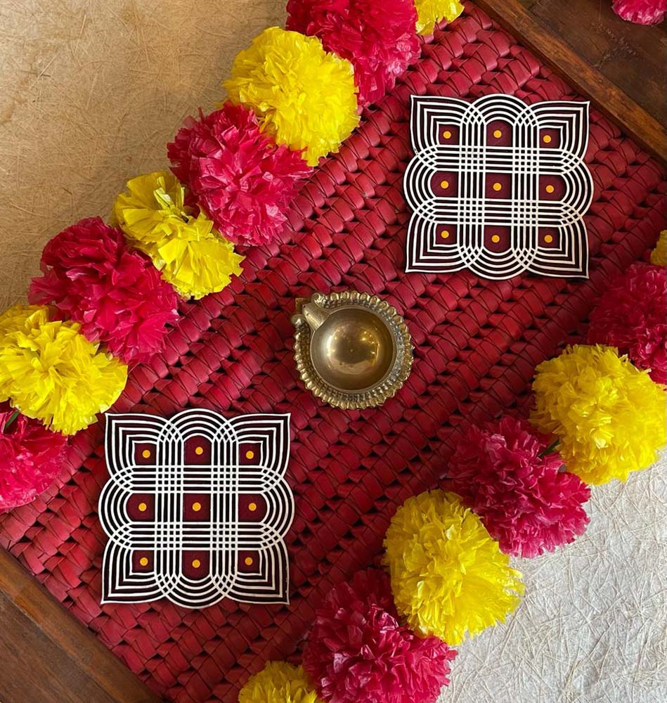 Set of 2 Margazhi Auspicious Kolams with intricate white designs and red dot accents, surrounded by yellow and pink floral garlands and a brass lamp.