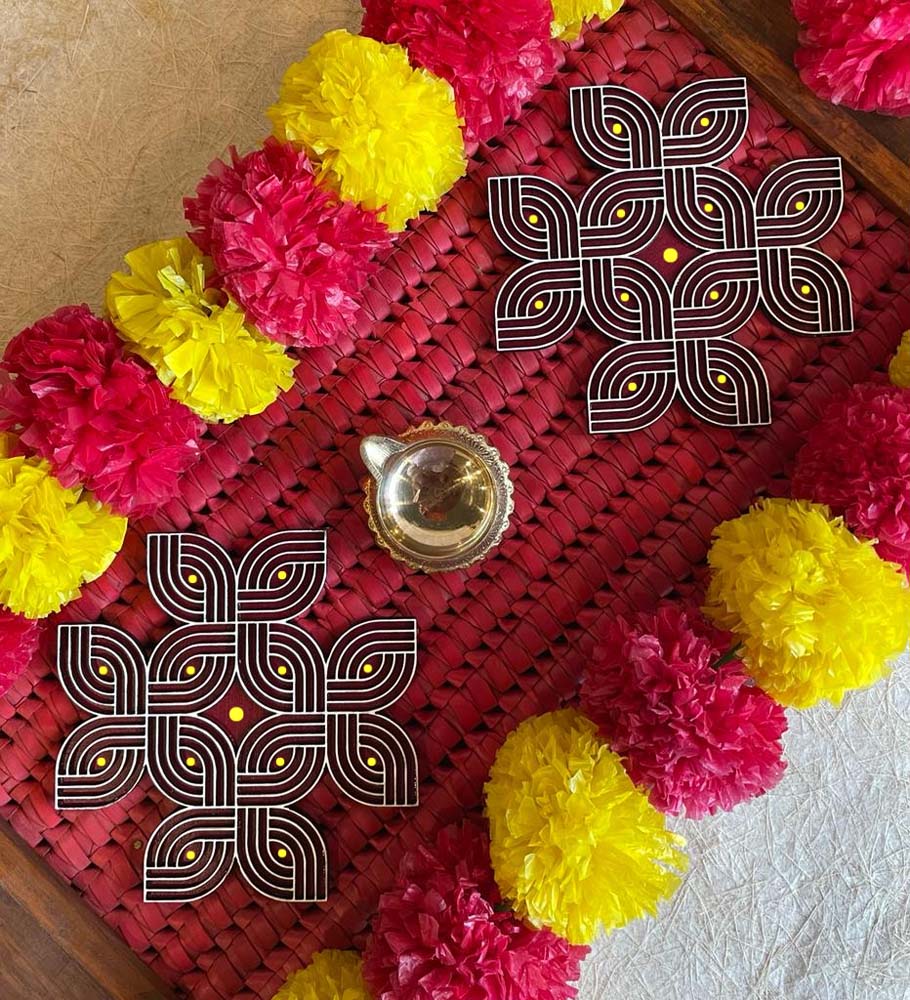 Set of 2 Margazhi-style Kolams with intricate dot patterns, placed on a red mat decorated with yellow and pink flowers and a brass lamp.
