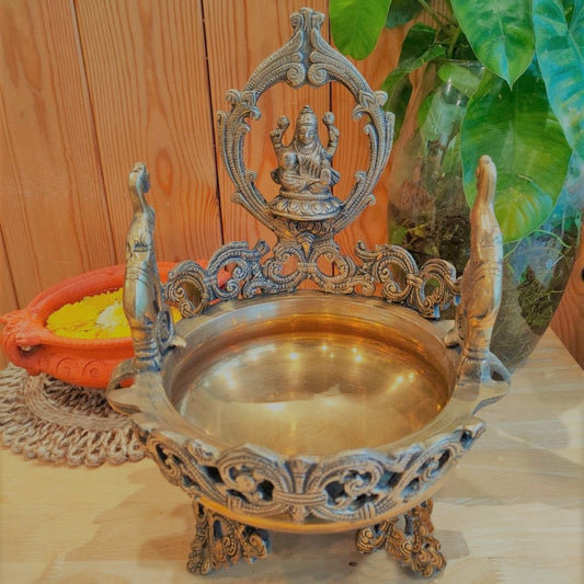 Intricately designed brass Lakshmi Urli with a central Goddess Lakshmi figure and peacock-shaped handles, displayed on a wooden surface with a terracotta pot and yellow flowers in the background.