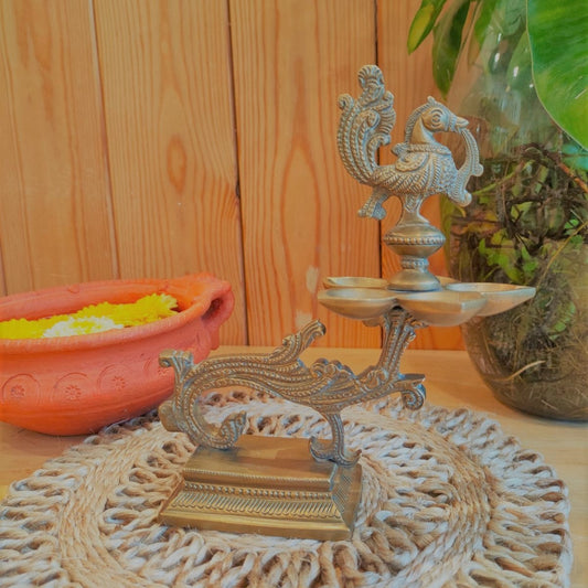 Traditional brass Peacock Lamp with an intricately designed peacock on top, mounted on a detailed Peedam, displayed on a wooden surface alongside a terracotta pot with yellow marigold flowers.