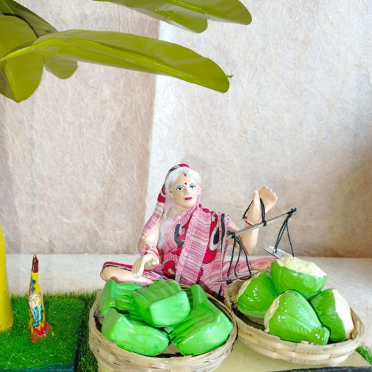 Navarathri Golu doll set featuring a female fruit seller in a vibrant saree, holding a weighing scale with baskets of green fruits, a banana tree, and a village deity.
