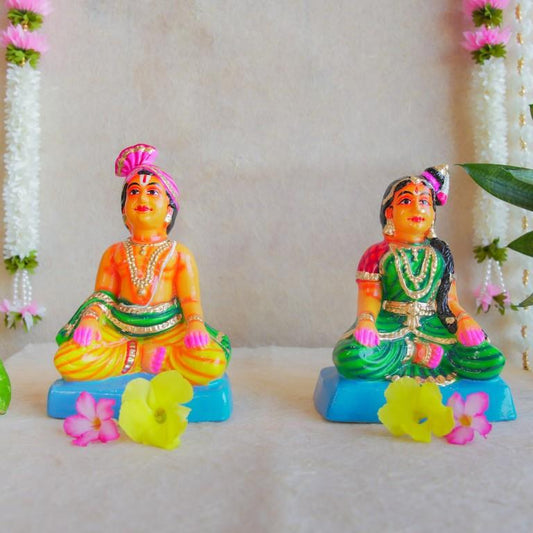 Navarathri Golu dolls depicting a seated Mama and Mami, dressed in traditional attire with vibrant colors, sitting on blue bases.