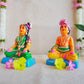 Navarathri Golu dolls depicting a seated Mama and Mami, dressed in traditional attire with vibrant colors, sitting on blue bases.