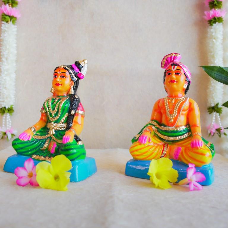 Navarathri Golu dolls depicting a seated Mama and Mami, dressed in traditional attire with vibrant colors, sitting on blue bases.