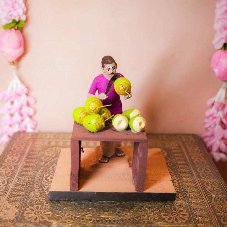 Handcrafted Golu Doll depicting a traditional coconut seller with fresh coconuts on a table, designed for Navarathri Golu.