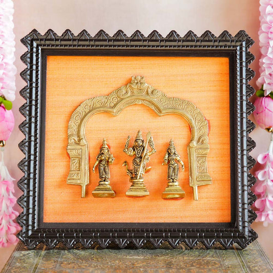 Framed brass sculpture of Lord Murugan with Valli and Deivanai under a Prabhavali, mounted on an orange fabric background with a carved wooden frame.