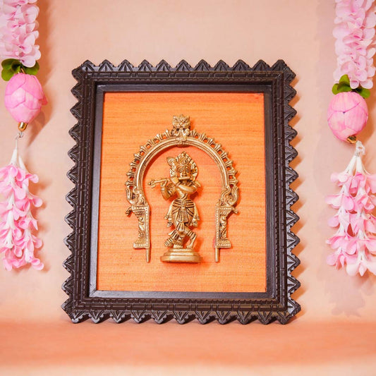 Framed brass sculpture of Lord Krishna playing the flute under an ornate arch, mounted on an orange background with a carved wooden frame.