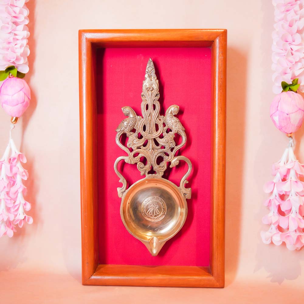 Framed brass Aarti lamp with intricate parrot design, mounted on a vibrant red fabric background with a polished wooden frame.