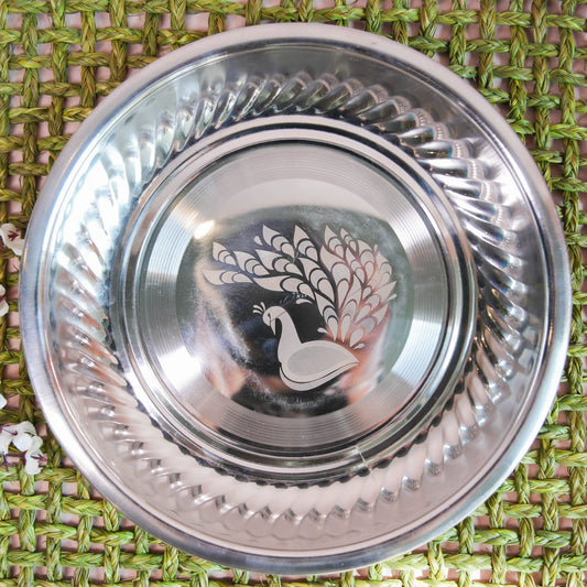 Stainless steel plate with an etched peacock design in the center and a contoured rim, placed on a green woven background with floral accents.