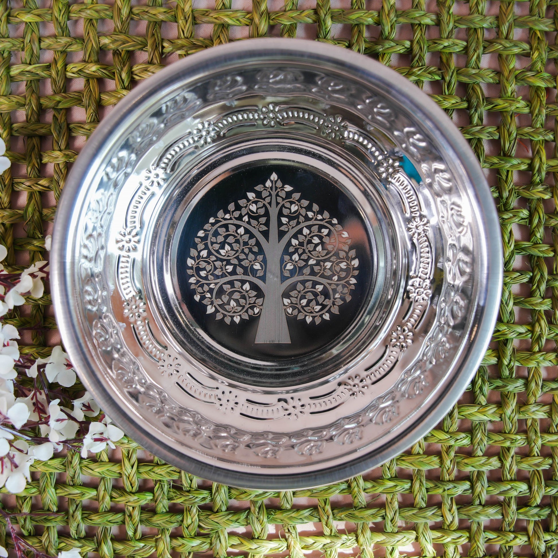 Stainless steel plate with a laser-etched tree design at the center, surrounded by intricate patterns, placed on a green woven background with floral accents.