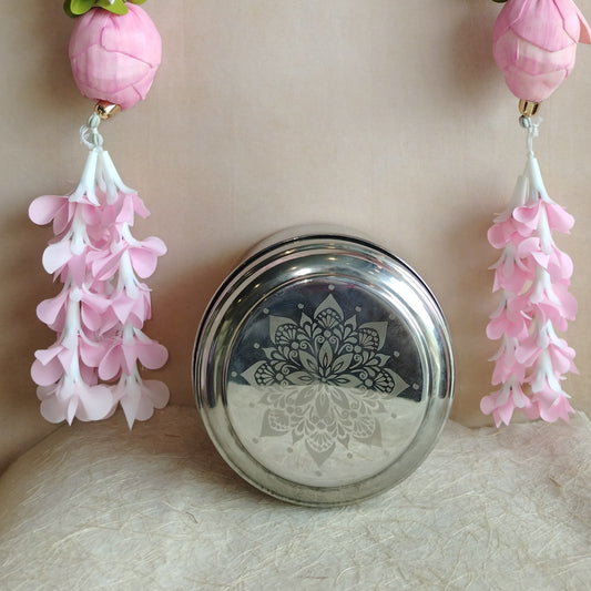 Stainless steel laddu box with a laser-etched floral design on the lid, placed against a decorative background with pink floral elements.