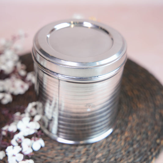 Small stainless steel storage box (Sambadam) with a polished finish, placed on a woven mat surrounded by floral decor.
