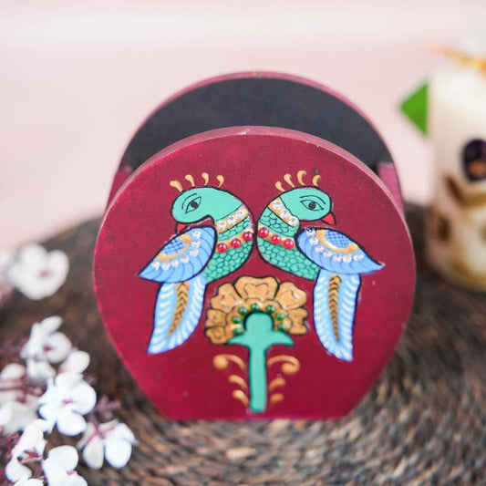 Hand-painted wooden penstand with a design of two colorful parrots perched above a floral motif, placed on a woven mat with decorative floral accents.