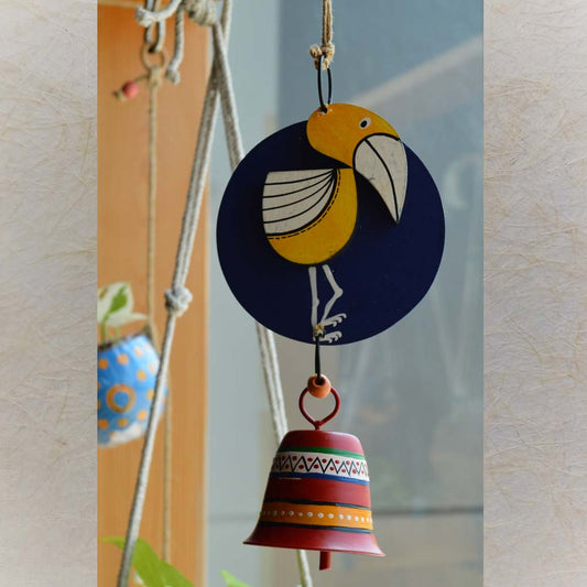 Hand-painted wooden yellow bird on a navy blue circular background with a colorful metal bell wind chime, hanging with rustic jute rope and wooden beads near a window and plants.