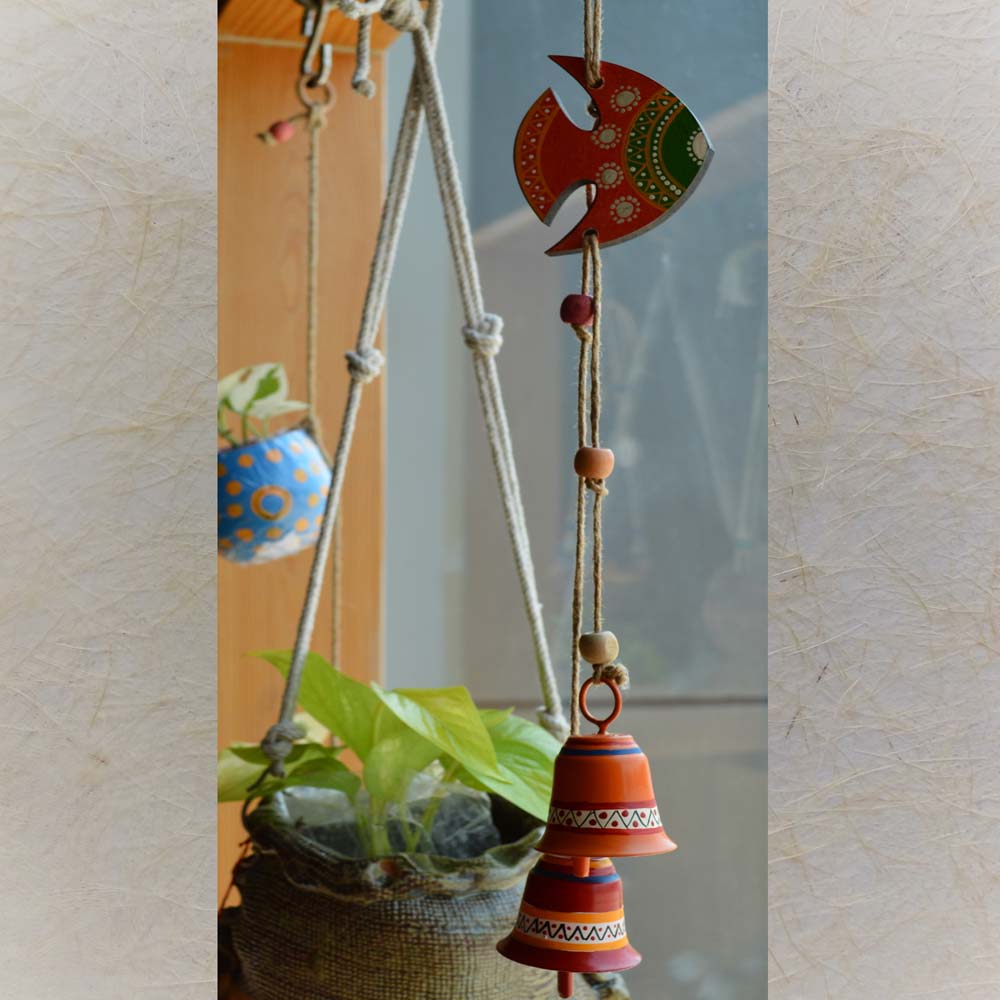 Hand-painted wooden fish in vibrant red and green with two colorful metal bells, hanging on a rustic jute rope with wooden beads near a window and plants.