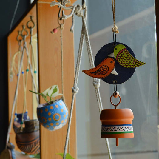 Hand-painted wooden love birds perched on a navy blue circular base with an orange metal bell wind chime, hanging with rustic jute rope and wooden beads near a window with plants.