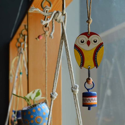 Hand-painted wooden owl in bright yellow with a colorful metal bell wind chime, hanging on rustic jute rope with wooden beads near a window and plants.
