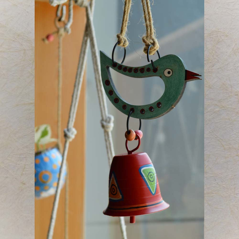 Hand-painted wooden bird in teal with a red metal bell wind chime, accented by jute rope and wooden beads, hanging near a window with plants in the background.