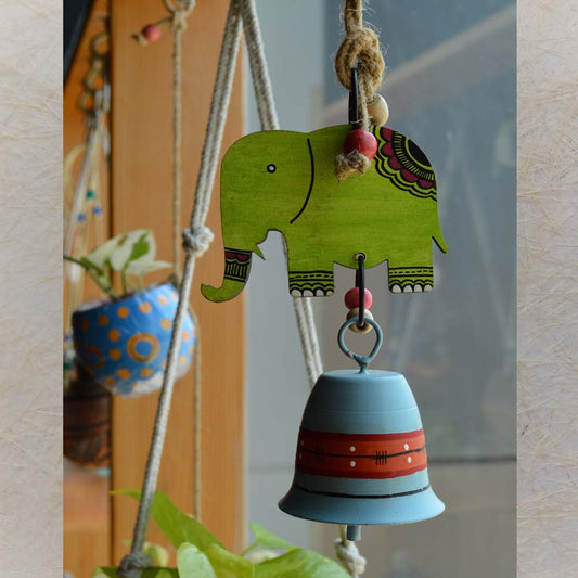 Wooden and metal hand-painted green elephant wind chime with a bell, adorned with jute rope and wooden beads, hanging near a window with plants.